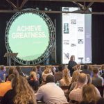 Picture of crowd from the side as they view an image on stage that says "achieve greatness"